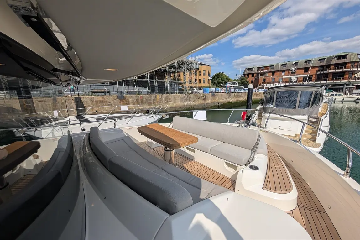 Luxury boat deck with grey seating and a wooden table, overlooking a tranquil marina and waterfront buildings.
